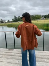 Blazer de gamuza sintética color terracota, con cierre frontal de botones y detalles de bordado a mano en contraste en los bordes, cuello y bolsillos. Presenta un diseño suave y flexible con mangas largas.