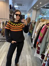 Sweater de punto tejido a mano en mohair, de color negro con diseño de guarda geométrica multicolor en tonos amarillo, naranja y rojo en la zona del canesú. Presenta cuello redondo y mangas largas con puños ajustados.