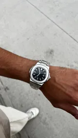Reloj de pulsera con caja de acero inoxidable de 40mm y esfera negra con textura horizontal. Presenta marcadores de hora luminiscentes, ventana de fecha a las tres y brazalete de acero inoxidable con acabado cepillado y pulido.
