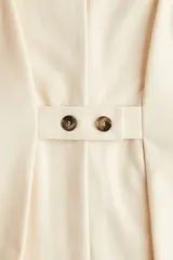 Chaleco largo color beige, con escote en V, cierre de botones a presión ocultos y bolsillos decorativos frontales. Presenta cintura entallada con pinzas y trabilla con botón en la espalda.