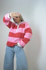 Sweater estilo rugby a rayas horizontales rosas y rojas con cuello blanco y cierre parcial con botones.