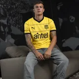 Camiseta de fútbol Puma del Club Peñarol, modelo Away Jersey. Diseño amarillo con detalles en negro en el cuello en V, los bordes de las mangas y líneas horizontales en el pecho. Incluye el logo de Puma, el escudo del club y el sponsor Antel.