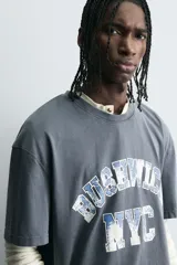 Camiseta de corte boxy fit con cuello redondo y manga corta. Presenta un diseño con efecto lavado y estampado gráfico frontal con tipografía universitaria.