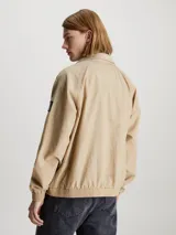 Campera bomber color beige de nailon reciclado, con cuello alto de canalé, puños y bajo elásticos, cierre de cremallera, bolsillos con botón de presión en la cintura, forro de poliéster reciclado e insignia con monograma de Calvin Klein en la manga.