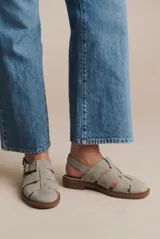 Sandalia tipo cangrejera de cuero color arena, con diseño de tiras anchas entrelazadas, talón descubierto y cierre mediante hebilla metálica lateral. Posee suela plana de goma color marrón con textura antideslizante.
