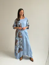 Vestido largo celeste con gran estampado floral marrón en el lateral derecho, de corte holgado, mangas 3/4 con trabilla y dobladillo irregular con flecos desflecados. Incluye un bolsillo aplicado en el frente.