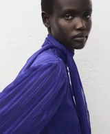Blusa de manga larga en color azul eléctrico con diseño de rayas verticales sutiles. Presenta un cuello alto con detalle de lazo integrado y cierre frontal con botones ocultos. El tejido es semitransparente con acabado satinado.