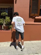 Remera blanca de manga corta con cuello redondo y estampado en la espalda con palmeras, ángel en una hamaca y las palabras 'Paradise Holiday' y 'Brixton'.