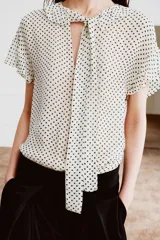 Blusa de viscosa con estampado de lunares, cuello redondo con escote en pico, volantes y lazada frontal. Presenta manga corta y bajo recto.