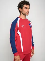 Buzo deportivo de manga larga, color principal rojo, con mangas azul marino y detalles en blanco que bajan desde el hombro hasta el puño. Presenta cuello redondo, logo Umbro y escudo del Club Nacional de Football en el pecho.