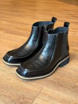 Botas cortas de cuero negro con textura de reptil, diseño de punta cuadrada y suela con detalle de franja en color beige.