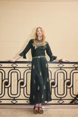 Vestido largo de satén color verde oscuro, con mangas tres cuartos terminadas en volados. Presenta un diseño con bordados dorados detallados en el pecho y cuello, además de motivos florales bordados dispersos a lo largo de la falda. Incluye un cinturón fino con aplicaciones brillantes.