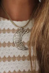 Collar con dije de luna creciente en metal plateado con diseño calado de líneas radiales, sostenido por un tiento de cuero fino en tono natural.
