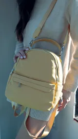 Mochila de tamaño mediano en color amarillo, confeccionada en símil cuero. Presenta un compartimento principal con cierre metálico, un bolsillo frontal con cierre y broche, y bolsillos laterales. Incluye correas regulables para la espalda, asa de mano y una correa adicional para llevar al hombro.