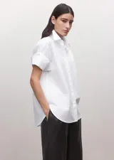 Camisa blanca de algodón con diseño recto, cuello camisero, manga corta y cierre frontal de botones ocultos con tapeta.