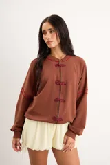 Campera marrón de corte oversize y largo intermedio con cierre frontal, detalles de broches de nudo estilo oriental y costuras bordadas en tono rojizo en cuello y mangas.
