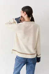 Buzo de punto color crudo con mangas de textura trenzada, detalles en contraste en los puños y franjas horizontales en el ruedo. Presenta cuello redondo y un pequeño bordado en el pecho.