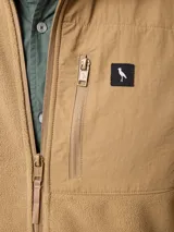Campera de polar color beige con cierre frontal completo y cuello alto. Presenta un bolsillo en el pecho con cierre y detalle de parche con logo. El diseño combina paneles de tejido polar con detalles en tejido técnico en la zona del pecho.