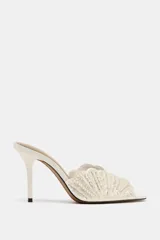 Sandalia de tacón alto estilo mule, de color blanco crudo, con diseño en la pala en forma de concha decorada con perlas y cuentas brillantes, punta cuadrada y tacón fino.