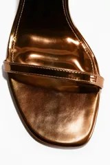 Sandalias de tacón alto fino con acabado metalizado dorado, diseño de tiras finas y tira ajustable al tobillo con hebilla.