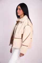 Campera de corderito color beige con ribetes y detalles en símil cuero marrón, cuello alto con hebilla decorativa, bolsillos laterales y cierre frontal.