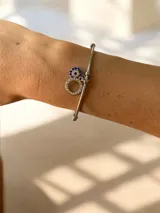 Pulsera tipo esclava de acero quirúrgico color dorado, con extremos esféricos y dijes colgantes: un círculo con strass y un dije de ojo turco con piedras azules y strass.