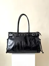 Bolso grande de cuero marrón con textura tipo cocodrilo, estructura rígida y asas para llevar al hombro. Tiene una tira con tachas y hebillas en cada extremo, también lleva lazos en los costados.