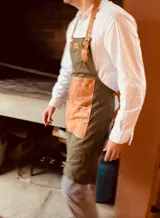 Delantal de cocina color verde musgo con detalles en cuero color suela. Tiene tiras de cuero ajustables al cuello y a la cintura, y dos bolsillos frontales de cuero. Ideal para asados y cocina en general.