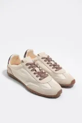 Zapatillas deportivas de color beige y crudo, confeccionadas en serraje vacuno con inserciones de tela, cordones marrones y suela de caucho con tacos. Cuenta con detalle de piel marrón en la parte trasera con logo.