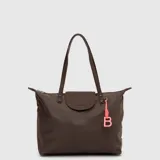 Tote bag color marrón, confeccionada en material impermeable, con doble asa de hombro, cierre superior y solapa frontal con logo grabado. Incluye un charm decorativo colgante con forma de letra B en color rosado.