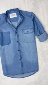 Camisa de jean celeste con cuello mao, manga larga con puños enrollados, cierre con botones y dos bolsillos frontales, uno de parche y otro con solapa y botón.