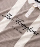 Remera tipo polo de corte oversized, confeccionada en tejido de punto con diseño de rayas verticales en tonos beige y blanco. Presenta cuello clásico, abertura frontal con botones y bordado con la inscripción 'The Hamptons New York' en el pecho.