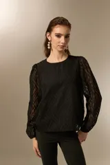 Blusa negra de poliéster con diseño texturizado de ondas, cuello redondo a la base y mangas largas transparentes con puños elásticos.