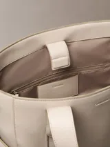 Cartera tote Calvin Klein de color beige, confeccionada en suave piel sintética, con diseño estructurado, asas fijas y compartimentos exteriores en los laterales. Presenta detalles de cinta con logo en jacquard y logo metálico grabado en la parte delantera.