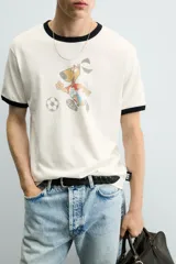 Camiseta de algodón color crudo con cuello y bordes de mangas en contraste negro. Presenta un estampado frontal de estilo vintage con el personaje Striker, mascota de la Copa Mundial de la FIFA 1994, jugando al fútbol.