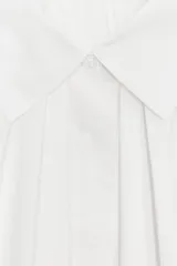 Camisa blanca de algodón con cuello solapa, detalle de pliegues en el delantero y mangas tres cuartos abullonadas terminadas en puño. Cierre frontal con botones ocultos.