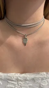 Gargantilla tipo choker compuesta por tiras de gamuza en color gris claro, con un dije metálico en forma de hoja.