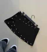 Minifalda negra de corte cruzado con detalle de tachas metálicas plateadas distribuidas a lo largo de los bordes y la superficie.
