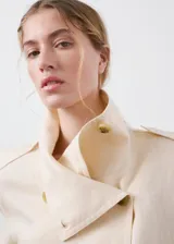 Chaqueta crop de lino color beige, diseño recto, cuello alto tipo embudo, manga larga, dos bolsillos laterales y cierre cruzado de doble botonadura metálica dorada con detalles en los hombros.