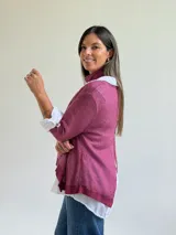 Sweater de punto color magenta con diseño asimétrico, cuello redondo y detalle de encaje bordado en el frente. Incluye una bufanda del mismo tejido y color, y se presenta superpuesto sobre una camisa blanca de algodón.