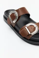 Sandalia tipo slide color tostado, con doble tira ancha y hebillas metálicas plateadas con diseño texturado. Su base es ergonómica y liviana, de color negro.