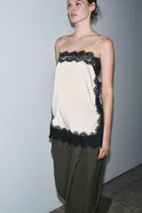 Top lencero de escote recto y tirantes finos, color crema, con aplicación de encaje negro contrastante en todo el contorno. Presenta aberturas laterales en el bajo.