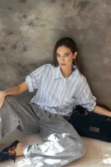 Camisa de corte recto y relajado, con cuello clásico, mangas cortas con detalle de ajuste y cierre frontal con botones. Presenta un diseño de rayas verticales en tonos celestes, blancos y grises.