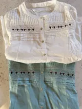 Blusa de manga larga color blanco con cuello redondo y cierre frontal de botones. Presenta detalles de bordado estilo punto smock en el pecho y textura de rayas verticales en la parte superior. Diseño holgado y estilo bohemio.