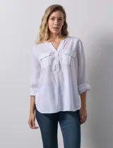 Blusa blanca de lino y rayón, con cuello a la base, escote en V y abertura con botones. Tiene bolsillos en la parte delantera y mangas largas regulables con presilla y botón.