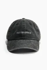 Gorra de sarga de algodón con efecto lavado, color negro, con la frase 'OUT OF OFFICE' bordada en blanco en el frente. Cuenta con trabilla ajustable y cierre de metal en la parte posterior.