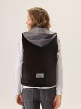 Chaleco gris con cuello alto y capucha, cierre frontal con botones a presión. La parte delantera es de lana gris lisa y la parte interior del cuello y el forro son de tela con patrón de espiga en blanco y negro.