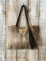 Cartera tipo tote de cuero vacuno con pelo, en tonos naturales jaspeados. Cuenta con manijas de cuero marrón oscuro, cierre frontal metálico dorado y un detalle de borla de cuero colgante.