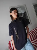 Remera tipo polo de corte recto y calce relajado, color azul marino, con cuello clásico, cartera frontal de botones y mangas cortas. Presenta bordados en contraste en el frente.