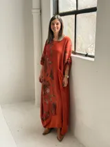 Vestido largo color terracota con diseño asimétrico que incluye un lateral con estampado floral grande. Presenta mangas tres cuartos, escote redondo y un bolsillo aplicado en el frente.